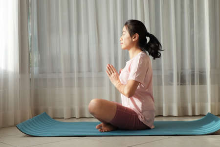 Young Asian woman lady practicing yoga alone in the prayer position at home window light.の写真素材
