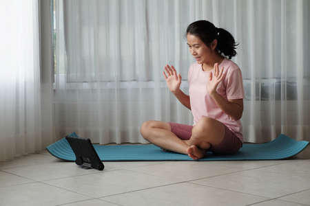 Young Asian woman says hello to the trainer teacher before starting a class online fitness workout at home window light. New normal Stay home concept idea.の写真素材