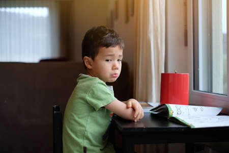 Portrait of Sad little kid boy sitting on the black chair next to the window and does not want to play, warm light tone.の写真素材