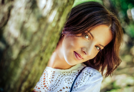 Close up Pretty Young Woman Peeking From Behind a Tree, Smiling at the Camera.の写真素材