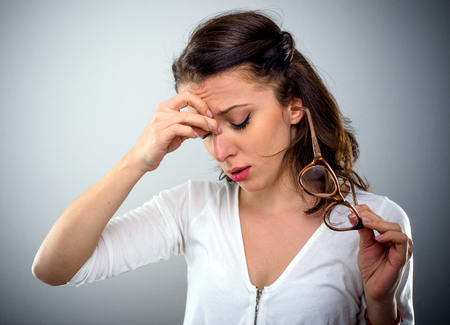 Attractive young woman with a headache holding her glasses in one hand as she pinches the bridge of her nose with a pained expression, head and shoulders on greyの写真素材