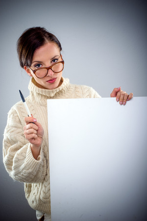 Scholarly attractive serious young woman holding a blank white sign as she peers over her glasses at the camera with a pen in her handの写真素材