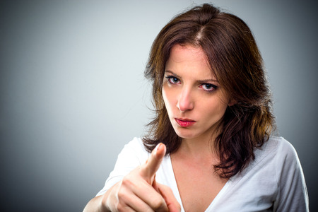 Young woman with a serious expression pointing a finger of blame and accusation, head and shoulders on grey with copy spaceの写真素材