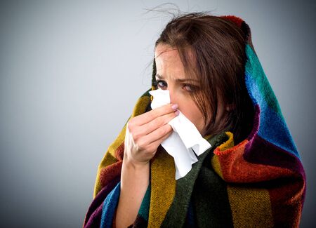 Young woman with a winter cold bundled up warmly on a colorful blanket blowing her nose on a tissueの写真素材
