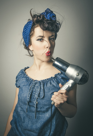 Half body portrait of housewife in vintage clothes pointing modern hair dryer at faceの写真素材