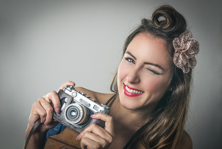 Fun vivacious retro fashion photographer with her hair in a French roll adorned with a flower winking at the camera with a beaming smileの写真素材