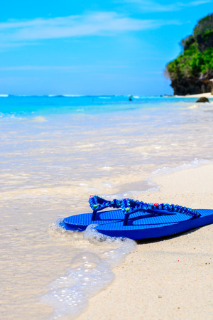 Wave washing up over pair of blue sandals on a tropical beach in Bali , Indonesia.の写真素材