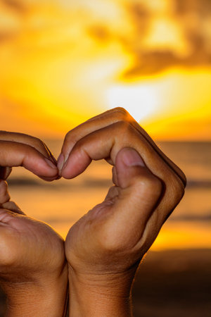 A woman making the love heart shape with her hands and a vivid orange tropical sunset in the...