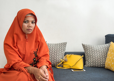 Asian woman wearing orange hijab outfit sitting on sofa looking at camera.の写真素材