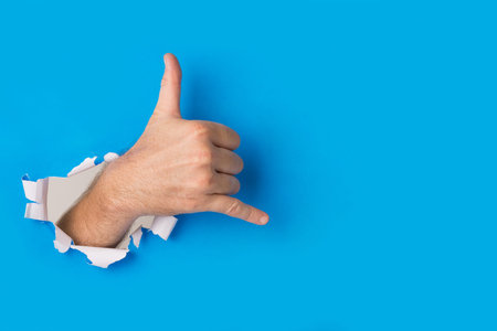 Male hand tearing through blue paper background creating a shaka hand gesture. A friendly hand sign...