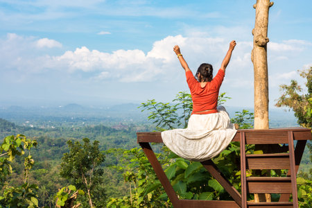Woman sitting on lookout raising her arms up into the air in a moment of happiness and success as she looks out at the view from a scenic view pointの写真素材