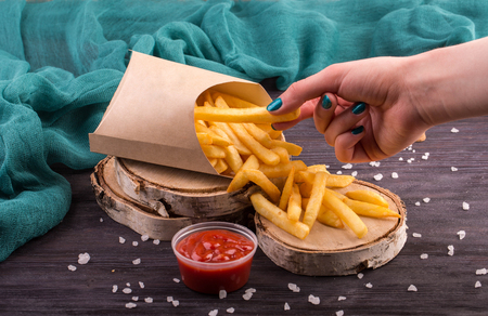 The female hand taking French fries from a cardboard box which lies on wooden saw cutsの写真素材