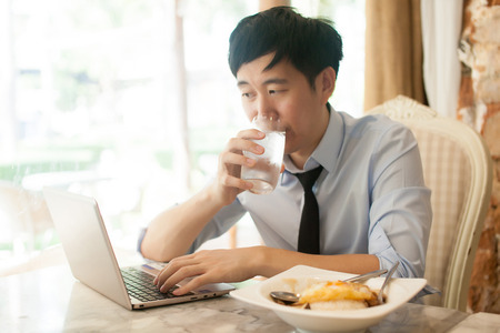 Young Asian man working while eating with his laptop in restaurantの写真素材