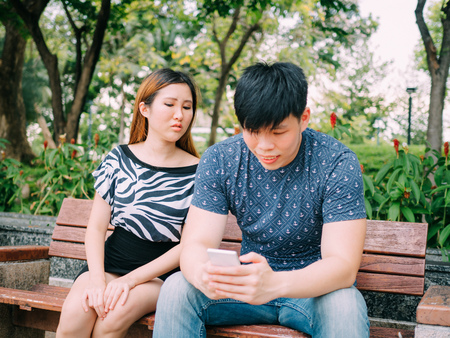 Jealous girlfriend peeking and spying her boyfriend mobile phone while he is reading a messageの写真素材