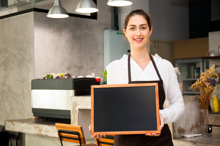 Beautiful Caucasian woman in barista apron holding empty blackboard sign inside coffee shop - ready to insert textの写真素材