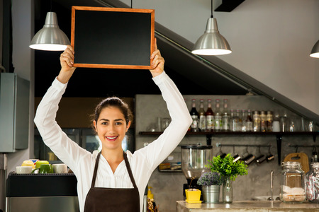 Beautiful Caucasian woman in barista apron holding empty blackboard sign inside coffee shop - ready to insert textの写真素材