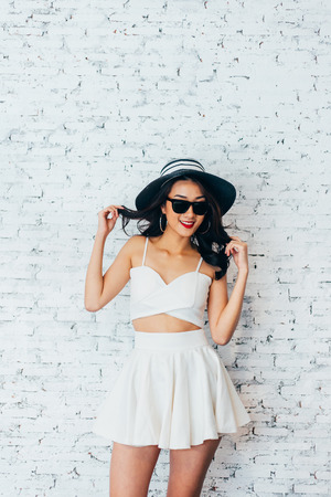 Young happy Asian woman smiling in fashionable dress and sunglasses and summer hat over white brick wall with copy spaceの写真素材
