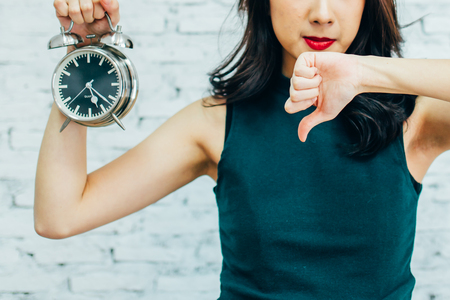 Asian business woman showing thumbs down and holding alarm clock - indicating that not pleased with time managementの写真素材