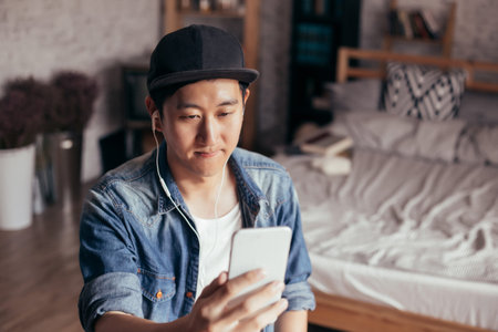 Young Asian man expressing serious look while talking video call via smartphone wearing headphones...