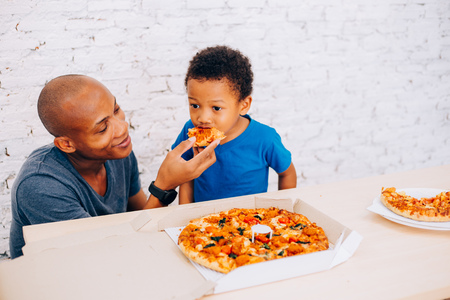 African American father feeding his cute son with pizza. Warm family concept. Warm family, Father and son relationshipの写真素材