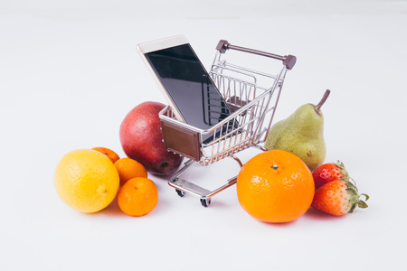 A shopping cart full of various kinds of fruits and groceries with smartphone inside, indicating mobile commerce and e-commerce for groceriesの写真素材
