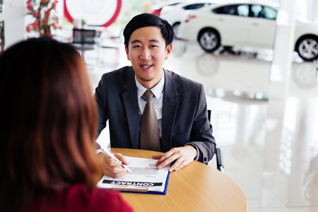 Young cheerful Asian business salesman giving new buyer a contract to sign for various purposes in car showroom - insurance, banking loan, and financingの写真素材