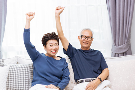 Senior Asian couple looking at camera and raising hands up while sitting on sofa at home