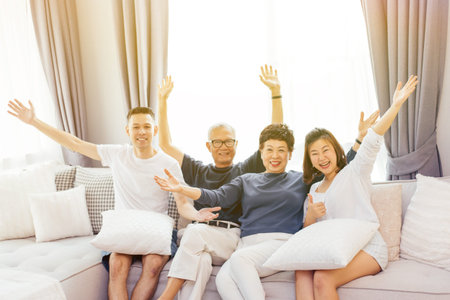 Asian family with adult children and senior parents raising hands up and sitting on a sofa at home....