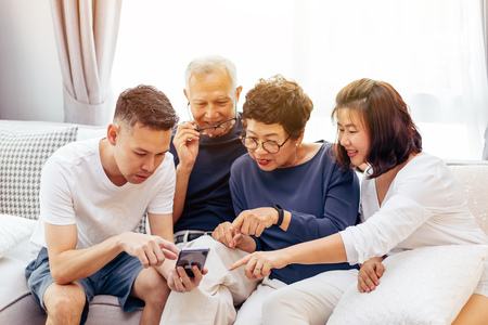 Asian family with adult children and senior parents using a mobile phone and relaxing on a sofa at home togetherの写真素材