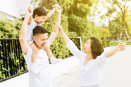 Cute Asian father piggybacking his son along with his wife in the park. Excited family raising hands together with happinessの写真素材