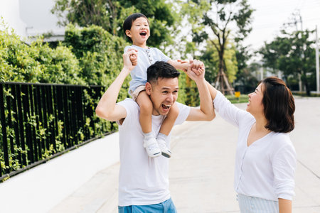 Cute Asian father piggybacking his son along with his wife in the park. Excited family raising hands...