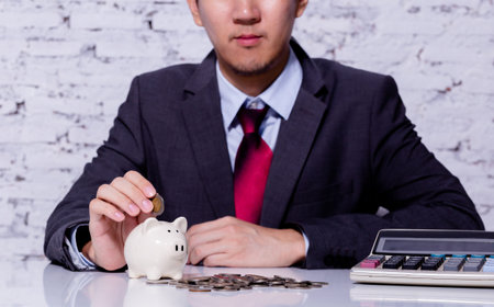 Businessman in formal suit putting and inserting coins in piggy bank with calculator nearby - business budgeting and saving for financial profit conceptの写真素材