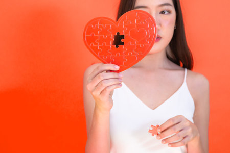 Portrait of attractive Asian woman holding red heart paper jigsaw isolated on pink red background