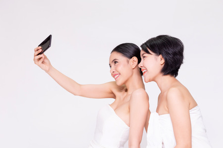 Young two Asian female best friends taking a selfie together with cellphone in white isolated backgroundの写真素材