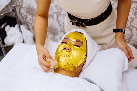 Beauty clinic staff helping young woman getting 24 karat gold facial treatment at the beauty clinic. The treatment of using real gold for youthful skinの写真素材