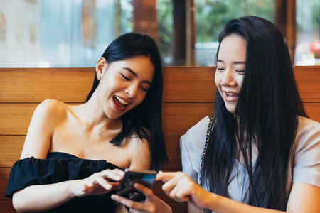 Two young attractive Asian women using a mobile phone together at restaurant with happiness and joyの写真素材