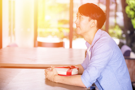 Young Asian man patiently sitting in cafe restaurant and holding a present gift giving to someone special for special occasionの写真素材
