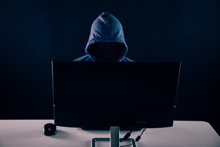 Anonymous and faceless hacker under hoodie using computer isolated over dark background - illegal...