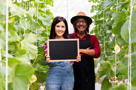 Young Asian beautiful woman holding an empty board with copy space alongside with senior South Asian man inside agricultural outdoors farming fieldの写真素材