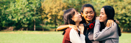 Portrait of multiracial and diverse all female teenage friends hugging and embracing together in the outdoor green park background - friendship and social happiness concept - banner sizeの写真素材