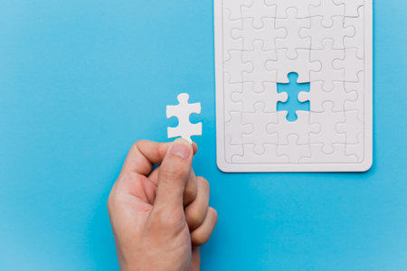Hands holding one last piece of jigsaw puzzle in blue background with unfinished white jigsaw puzzle...