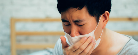 Young Asian man coughing and suffering in medical mask inside home bedroom - illness and fever concept - with copy space banner sizeの写真素材