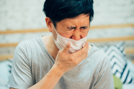 Young Asian man coughing and suffering in medical mask inside home bedroom - illness and fever concept