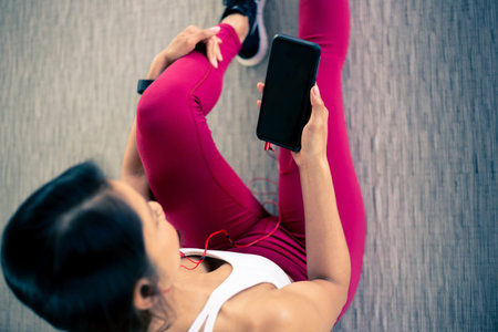 Top view - Young woman in sportswear using a smart phone and wearing earphones and listening inside...