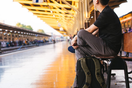 Young Asian male travel backpacker sitting and waiting for a train in railway platform in Bangkok,...