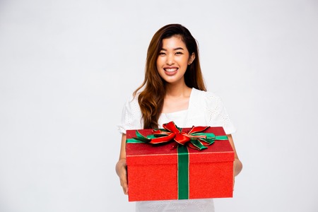 Lovely Asian female smiling and giving big gift box to camera while standing on gray backgroundの写真素材