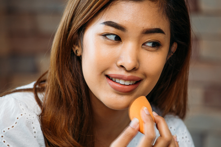 Closeup young Asian positive woman applying powder on shiny face on blurred backgroundの写真素材