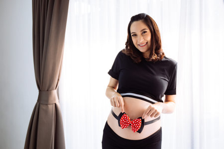 Young pregnant woman holding her tummy with a ribbon bow over the window view in the morning with...