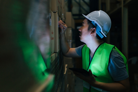 Side view of Asian young warehouse worker man with safety hard hat is checking order details with a digital tablet at inventory roomの写真素材