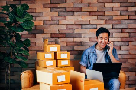 Young Asian male business entrepreneur use computer laptop and phone with boxes to deliver customers from orders - Online business and ecommerceの写真素材